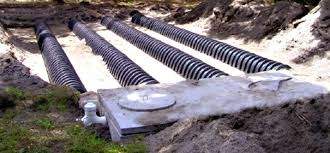 A septic system showing multiple black, corrugated pipes laid in trenches, connected to a concrete septic tank in a dug-out area of soil.